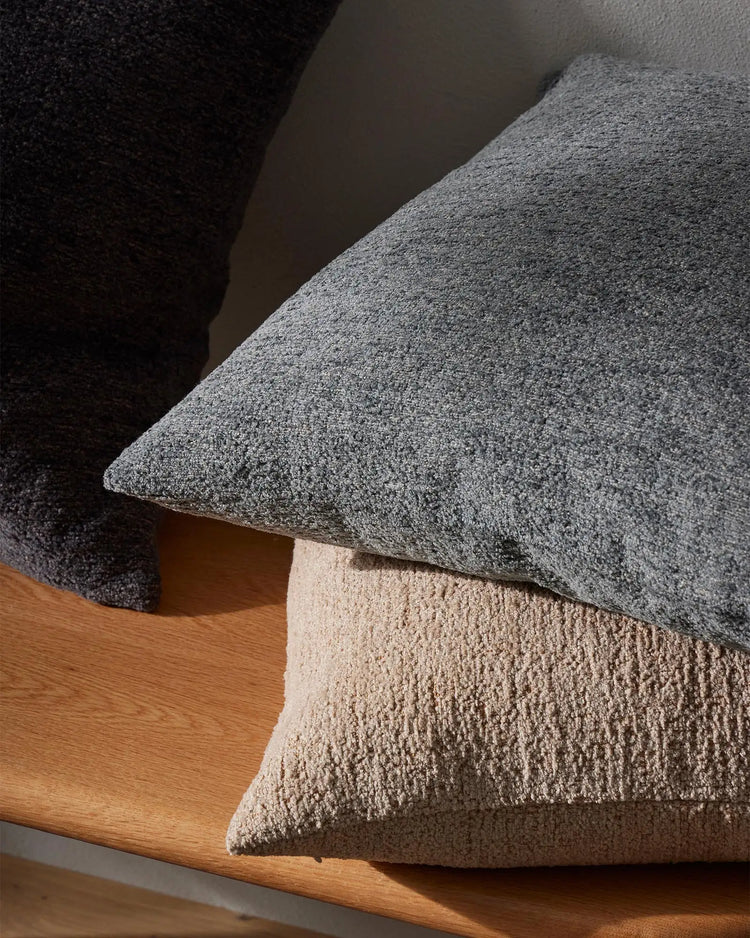 Textured cushion stack in charcoal and natural tones on wooden furniture, showcasing layered neutral palette and tactile woven fabrics in modern minimalist styling