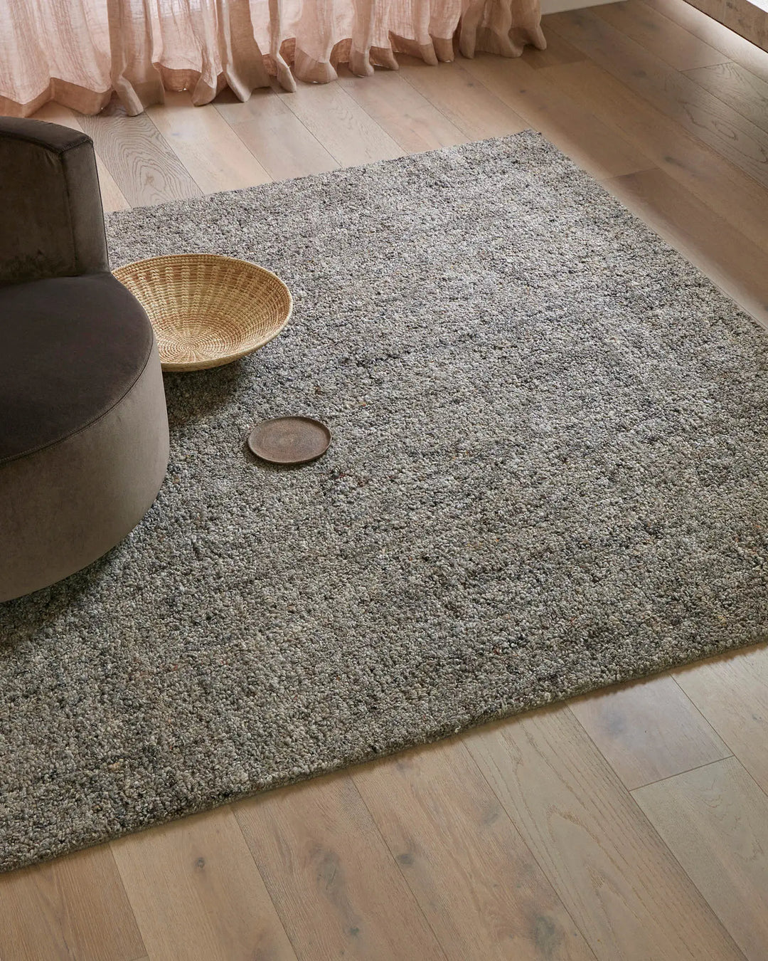 Minimalist living room with sage-grey wool blend rug layered on light wood flooring, styled with blush linen curtains, charcoal poufs and natural woven basket accessories