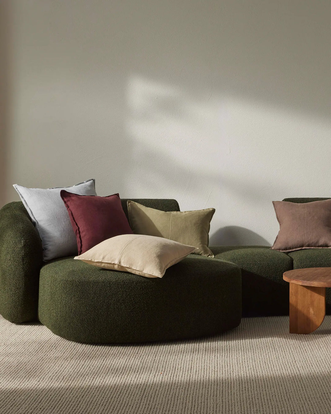 Earthy lounge styled with Como cushions in sandstone, mocha and willow tones layered on a forest-green curved sofa, soft natural light and neutral textured rug creating warm, organic modern aesthetic
