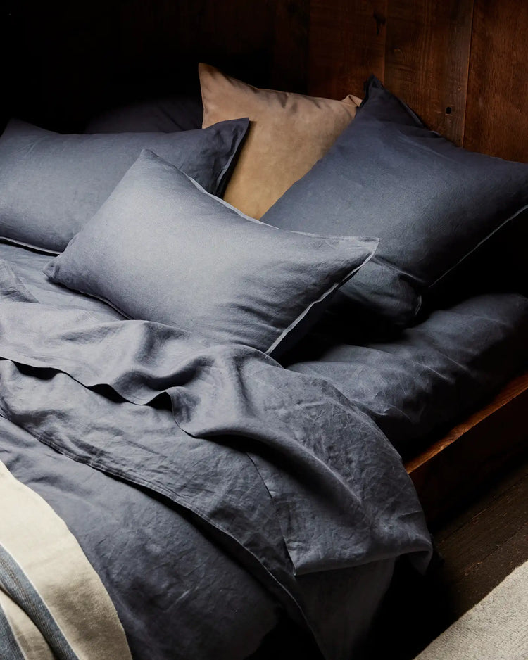 Rumpled denim linen bedding styled on wooden bed frame with layered pillows in cool blue-grey tones, showcasing textured European linen and relaxed, lived-in aesthetic