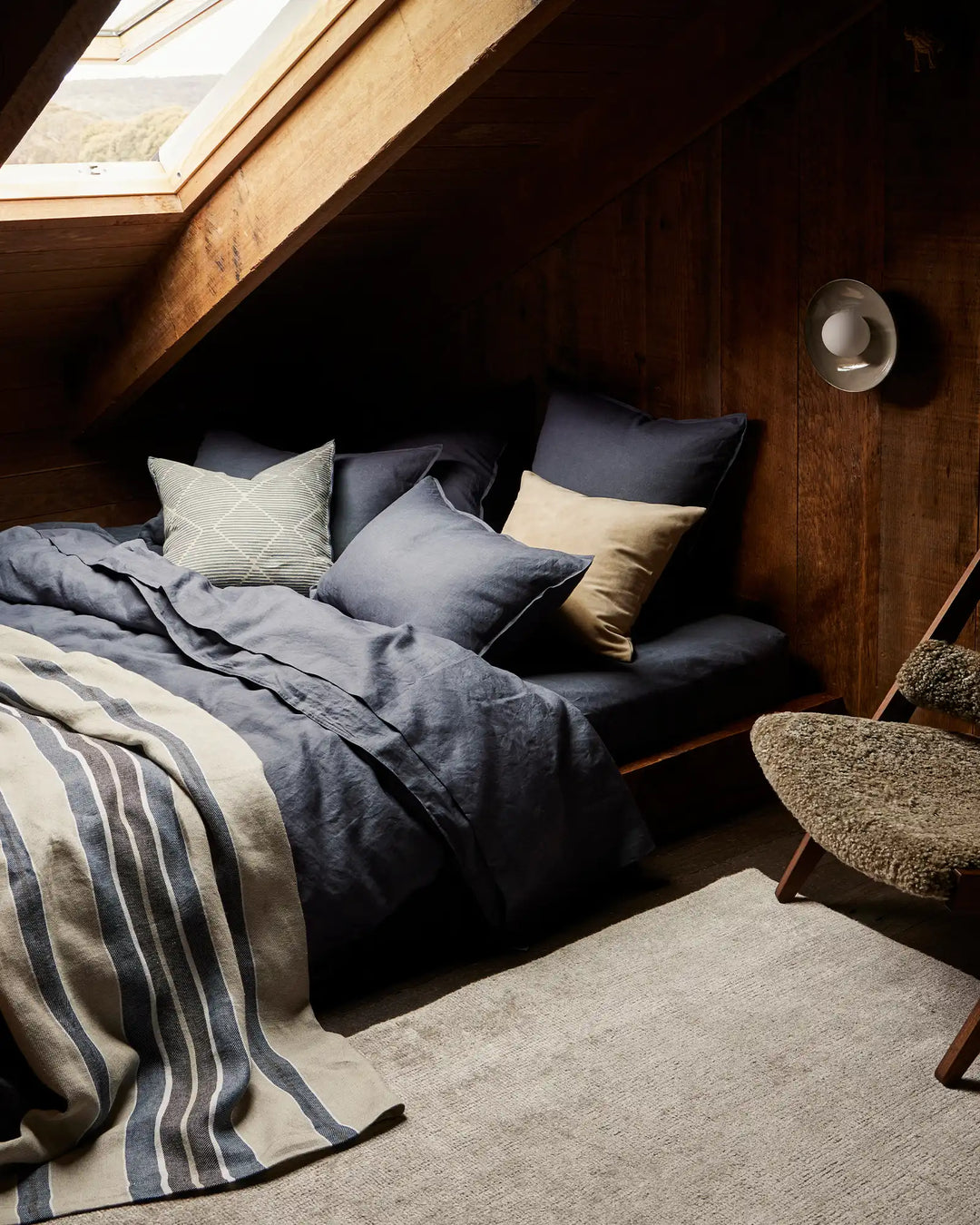 Attic bedroom with navy linen bedding and cream striped throw, pale geometric cushion, natural wood beams, skylight, and woven chair—coastal farmhouse aesthetic