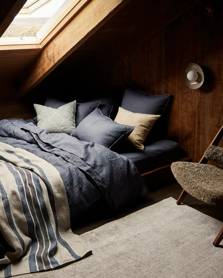 Attic bedroom with navy linen bedding and cream striped throw, pale geometric cushion, natural wood beams, skylight, and woven chair—coastal farmhouse aesthetic