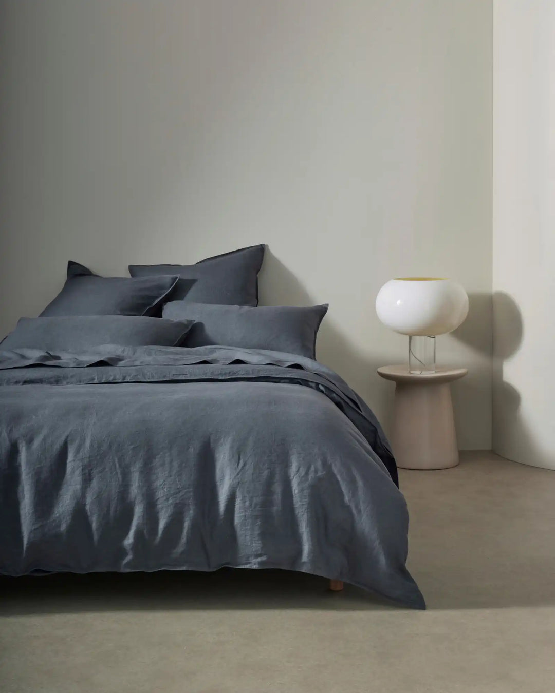 Minimalist bedroom styled with slate-blue linen bedding and layered pillows, soft neutral walls, timber nightstand, and sculptural white ceramic lamp creating calm contemporary aesthetic