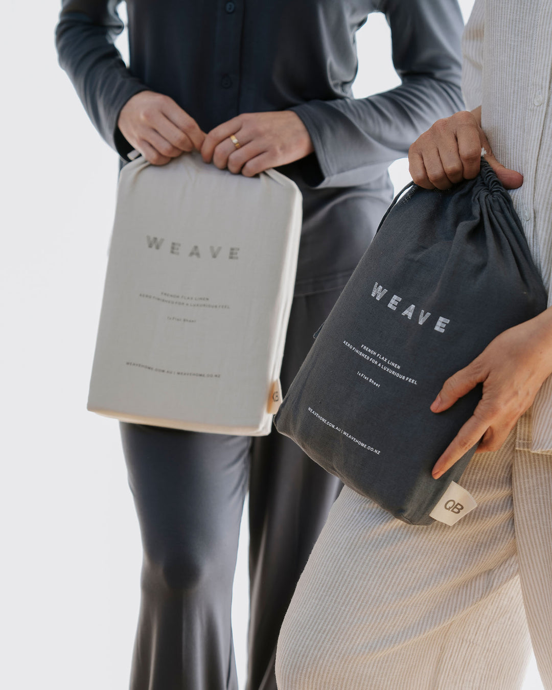 Two people holding packaged Ravello linen bedding in silver and denim, styled against neutral tones and textured fabrics in minimalist monochrome palette