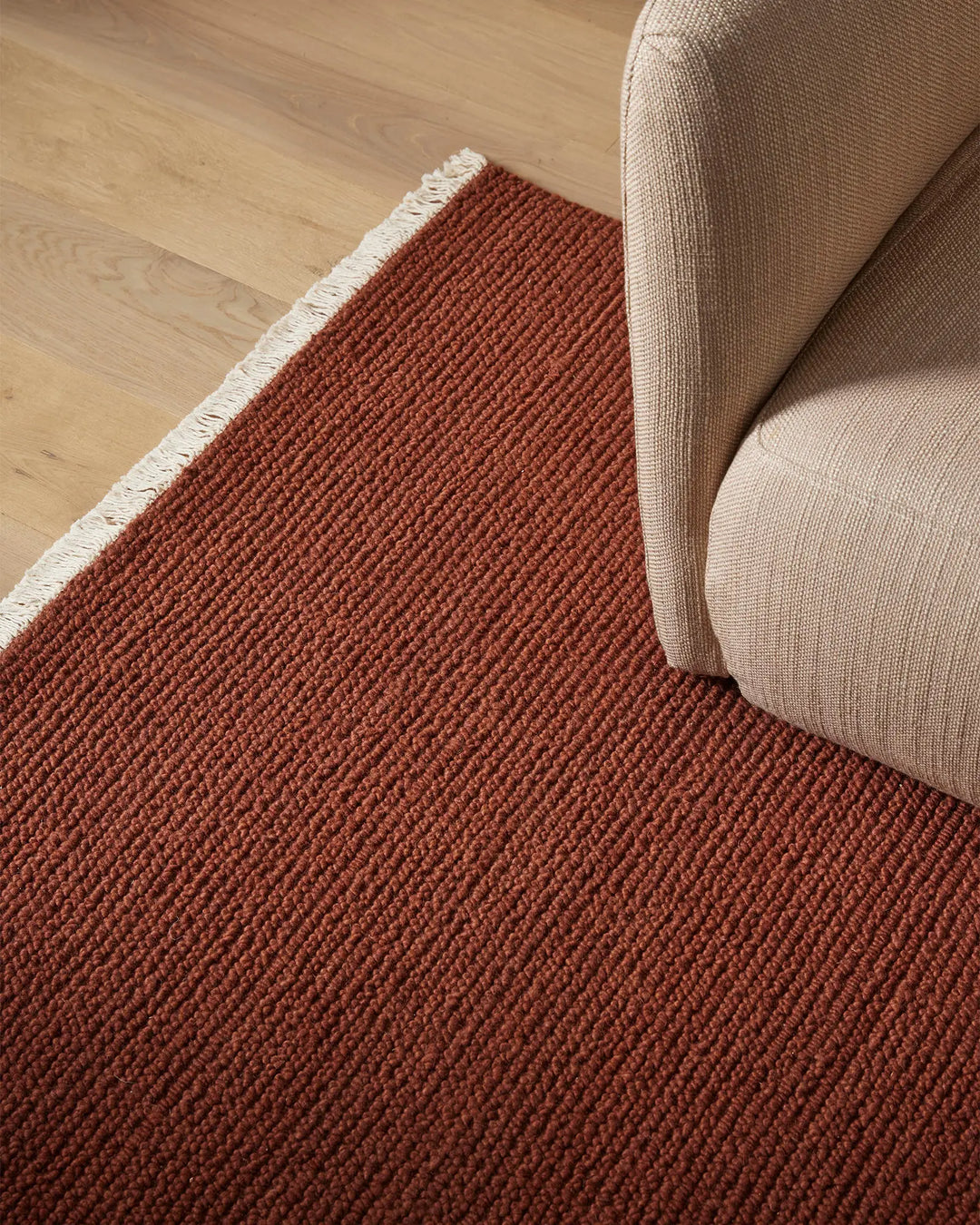 Warm terracotta wool rug with crisp white fringe beside natural beige upholstered furniture on light wood floor, earthy minimalist interior styling
