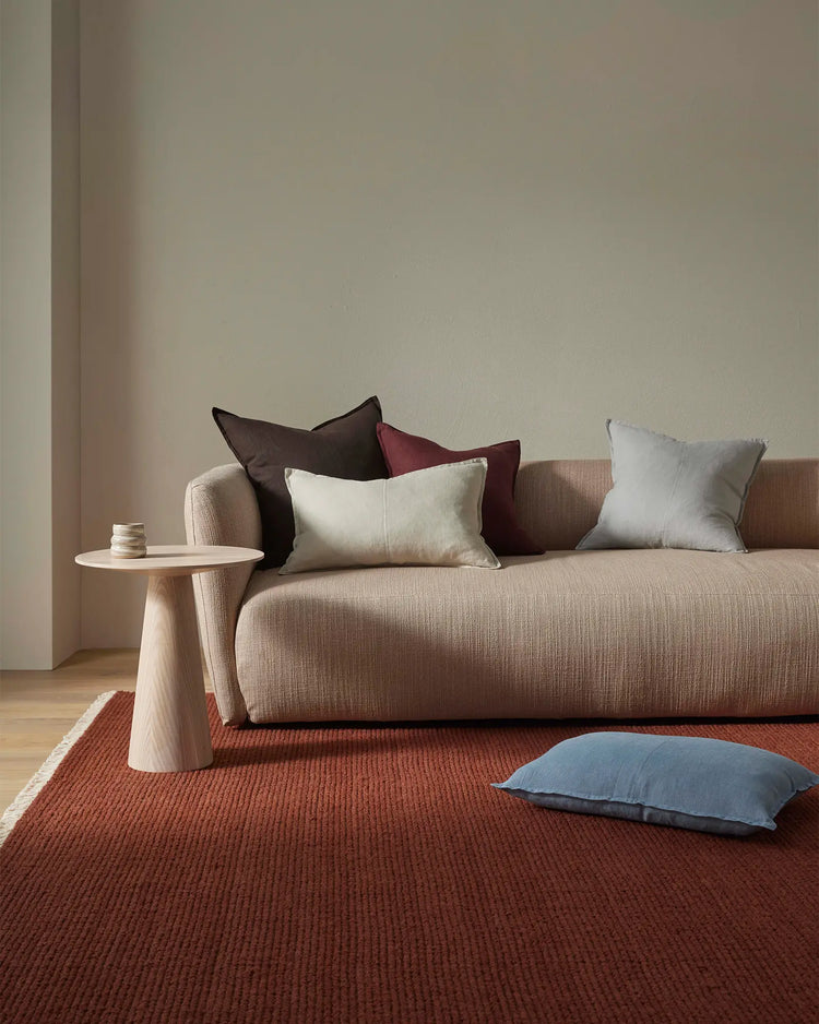 Warm minimalist living room with terracotta wool rug, beige upholstered sofa, and layered cushions in cream, burgundy and soft blue tones against neutral walls