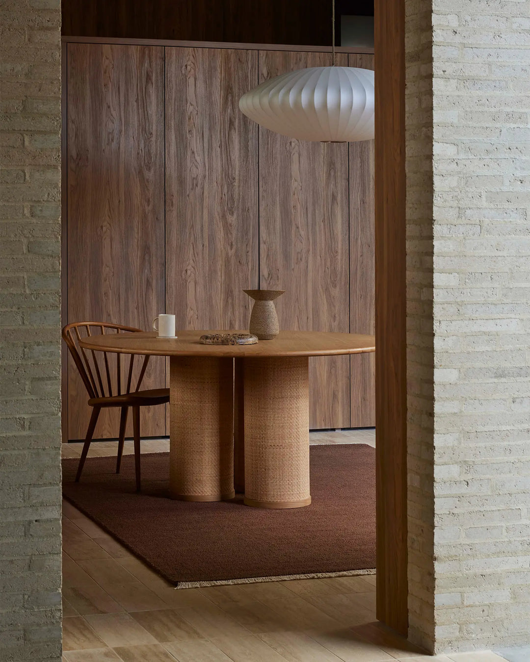Warm minimalist dining nook with textured wool rug in ember tones, natural wood table, woven cylindrical base, and mid-century chair against brick walls