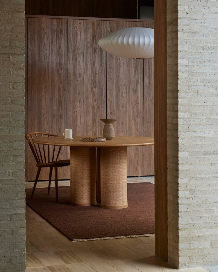 Warm minimalist dining nook with textured wool rug in ember tones, natural wood table, woven cylindrical base, and mid-century chair against brick walls