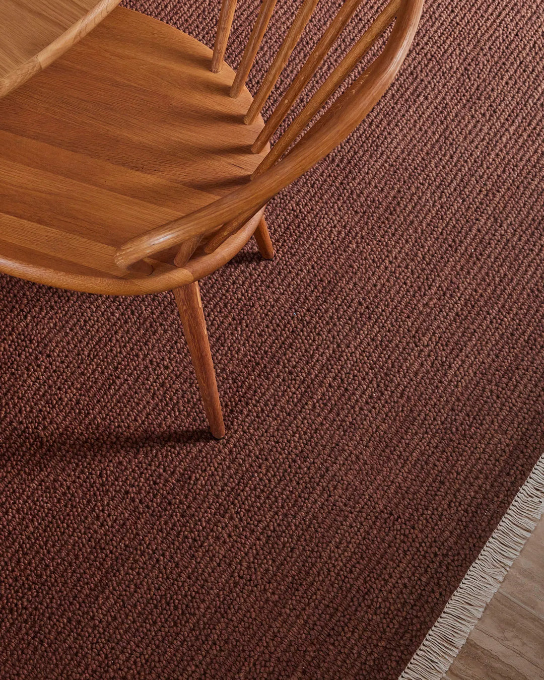 Warm terracotta wool rug with textured weave beneath mid-century wooden chair, styled in earthy, minimalist interior with natural wood tones