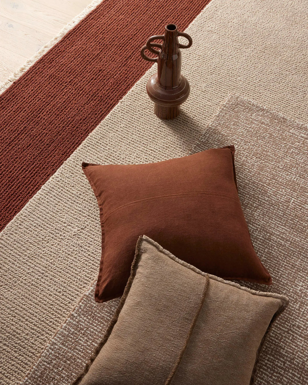 Warm terracotta and stone wool rugs layered with rust and taupe cushions, styled with sculptural timber accents in earthy, minimalist aesthetic