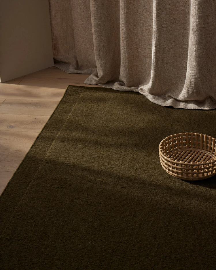 Earthy neutral interior with olive-toned wool rug, linen curtains in taupe, and woven basket accent on warm timber floor