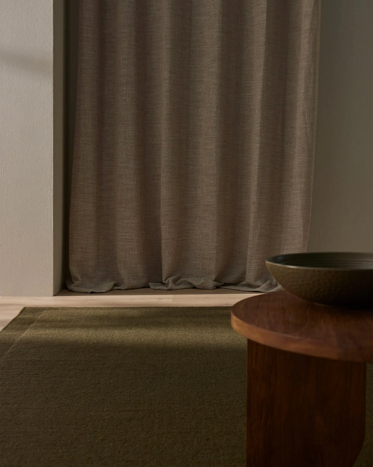 Earthy minimalist interior with sage-green wool rug, taupe linen curtains, and warm wood furnishings creating a calm, neutral palette