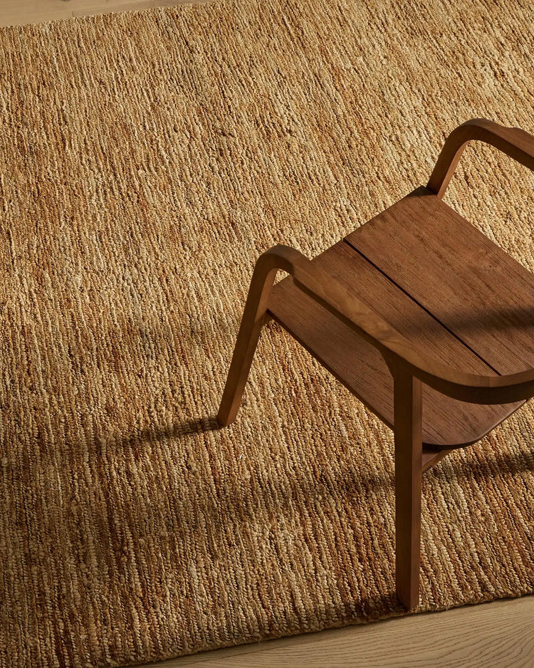 Warm natural jute rug with textured weave in a minimalist room setting, paired with mid-century wood chair casting soft shadows