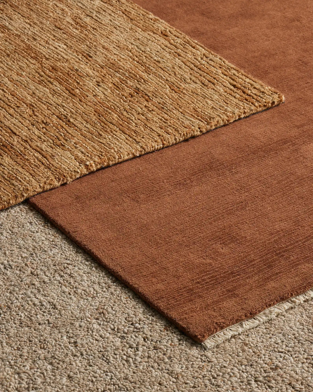 Warm terracotta and natural jute layered rugs with textured weaves in earthy tones, styled on soft cream carpeting for a warm, organic interior aesthetic