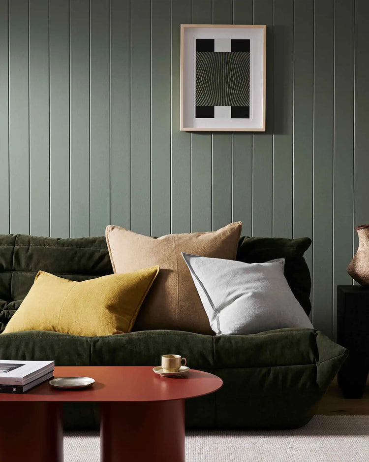 Sage green panelled living room with olive sofa styled with Como cushions in mustard, taupe, and white, geometric black artwork, terracotta accent table, minimal modern aesthetic