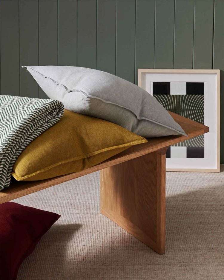 Styled bedroom vignette with stacked linen cushions in ochre and grey tones, textured throw, and timber bench against soft sage green panelled wall
