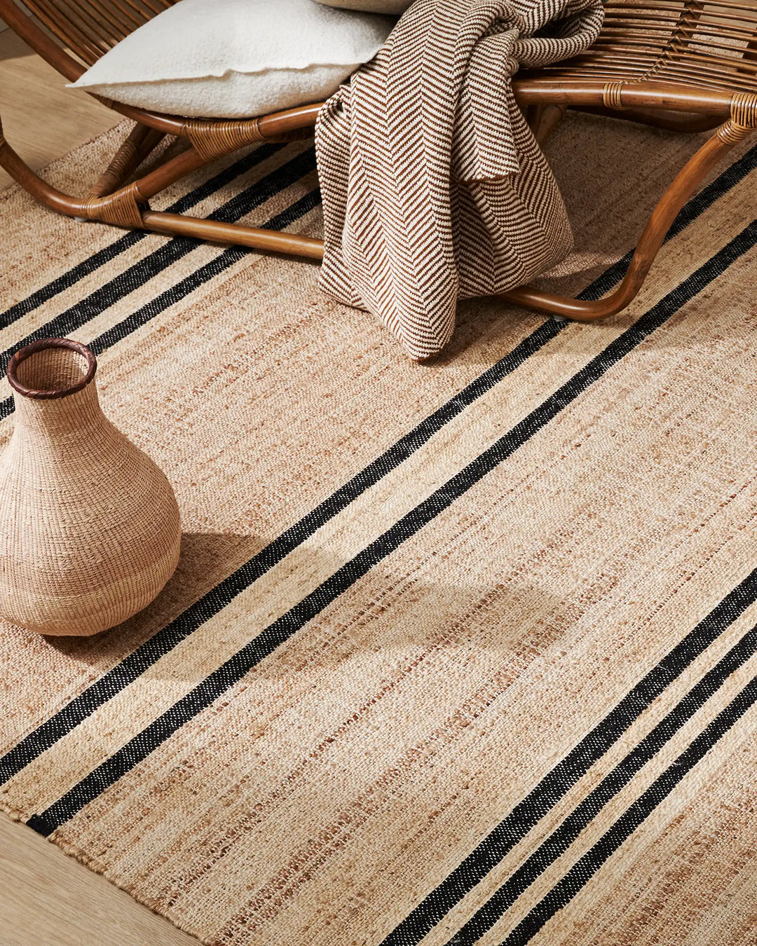 Neutral jute rug with black striping styled with rattan chair, herringbone throw, and woven vessel in earthy, organic living space