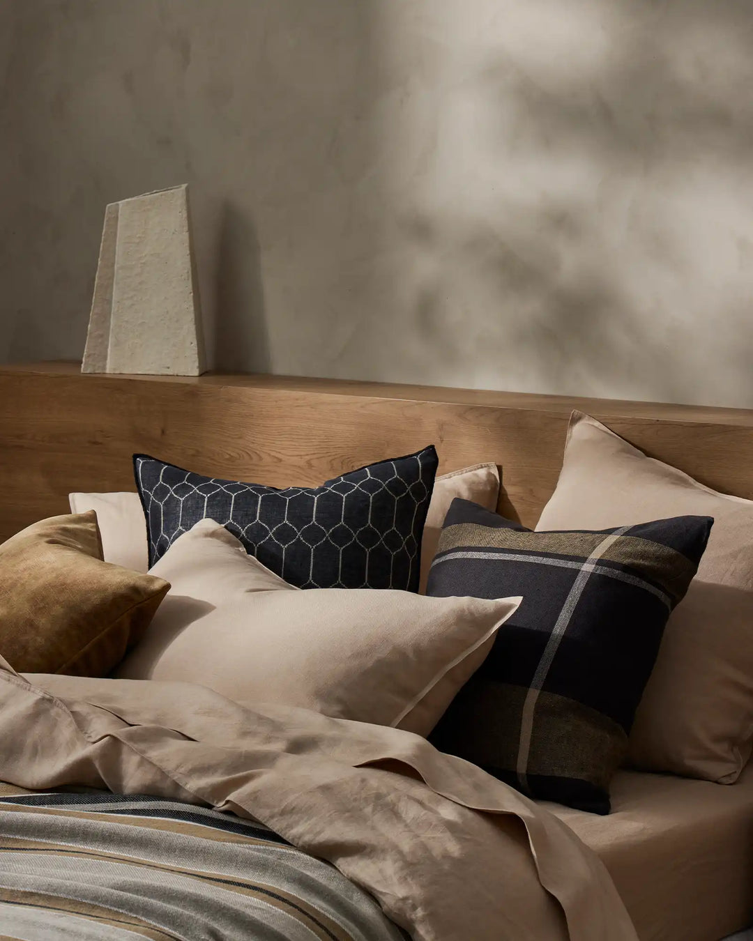 Neutral bedroom scene with layered cushions in cream, black and olive tones featuring the Syracuse and Dante ranges, soft linen textures, natural wood furniture and warm diffused light