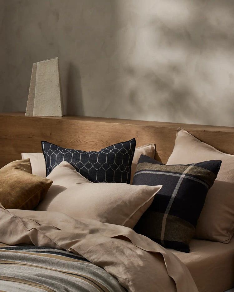 Neutral bedroom scene with layered cushions in cream, black and olive tones featuring the Syracuse and Dante ranges, soft linen textures, natural wood furniture and warm diffused light