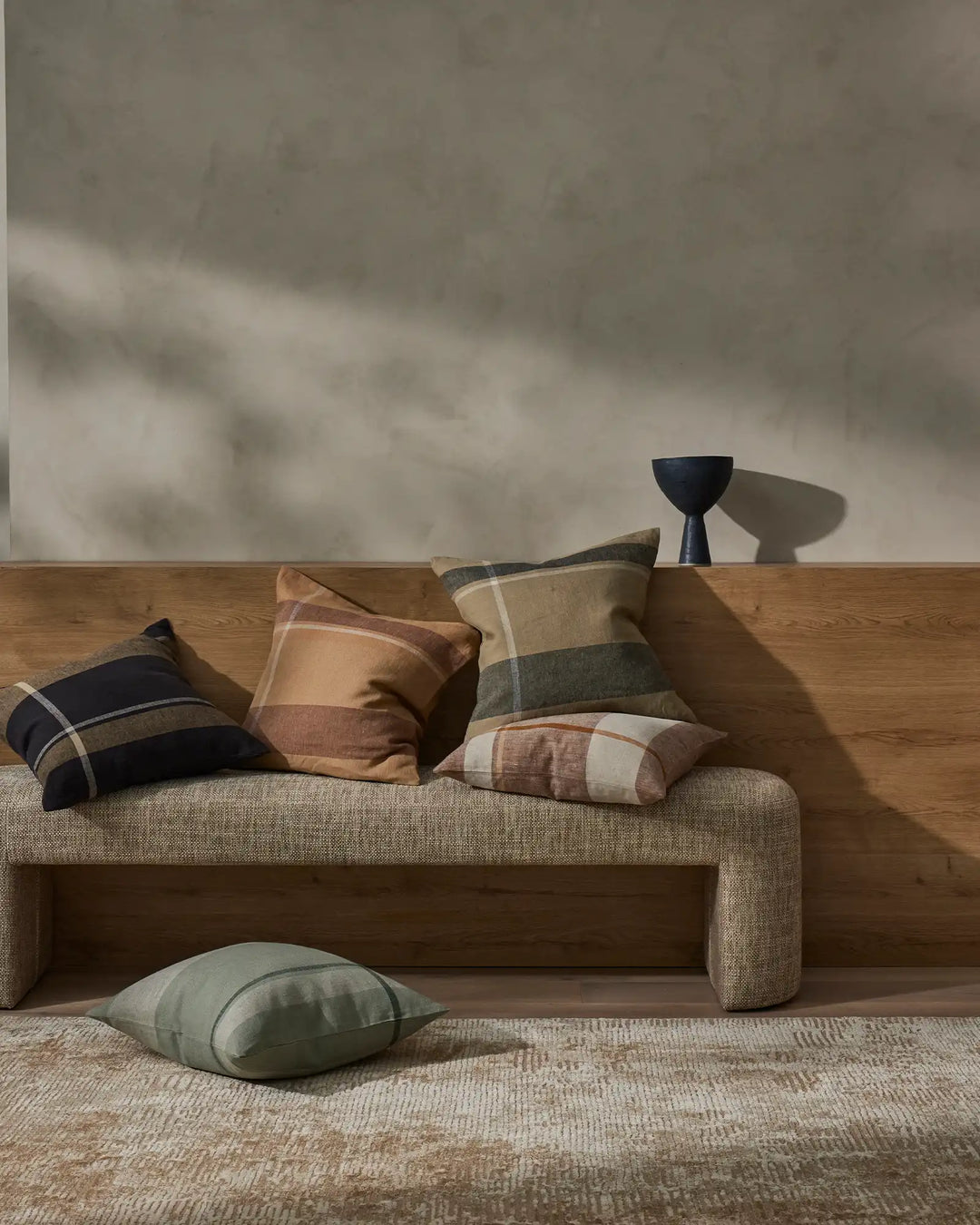 Warm earthy tones with navy accents: linen cushions in terracotta, tobacco and midnight checks arranged on a natural upholstered sofa, minimalist styling with soft shadow play on neutral walls