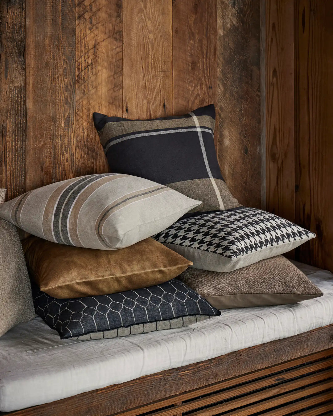 Layered neutral and charcoal cushions with geometric patterns and striping on weathered timber bed, earthy and modern Nordic-inspired bedroom styling
