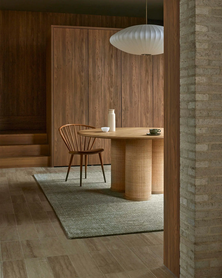 Mid-century modern entryway with sage green wool rug, warm timber furniture, and rattan table base against exposed brick and wooden paneling