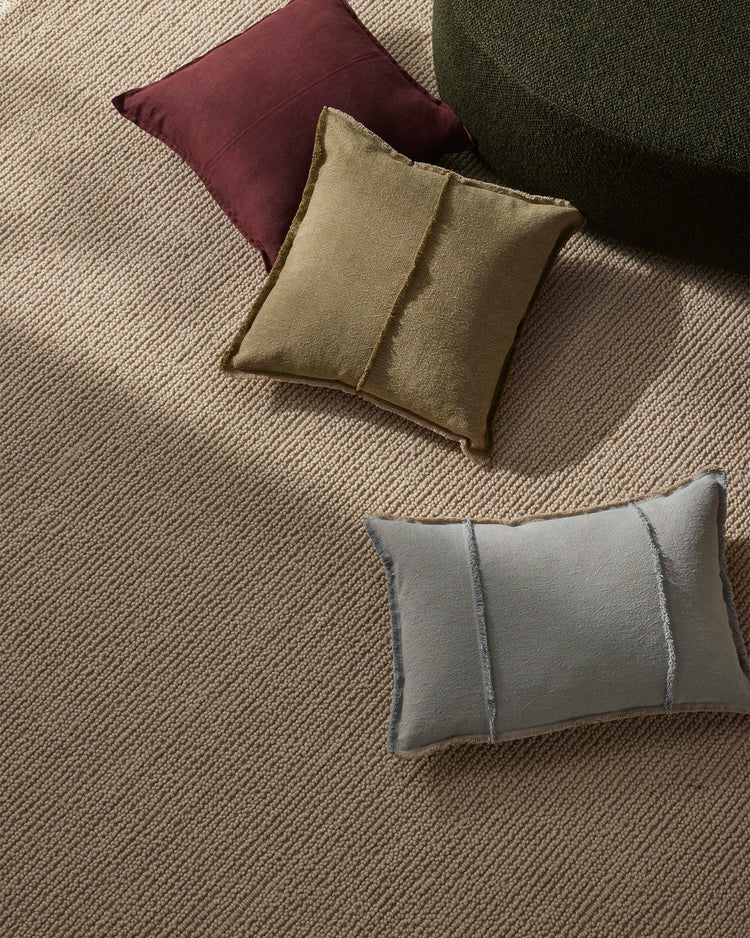 Overhead view of earth-toned cushions in burgundy, sage green, olive and stone grey arranged on a natural wool rug with subtle ribbed texture, creating a warm, layered interior styling palette