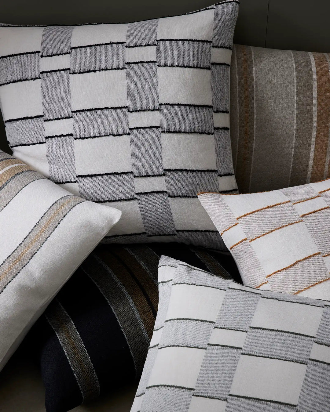 Neutral and earthy bedding styled together: geometric patterned cushions in cream, grey and charcoal with textured stripe details, layered with tonal striped pillows in warm bronze, taupe and cream tones