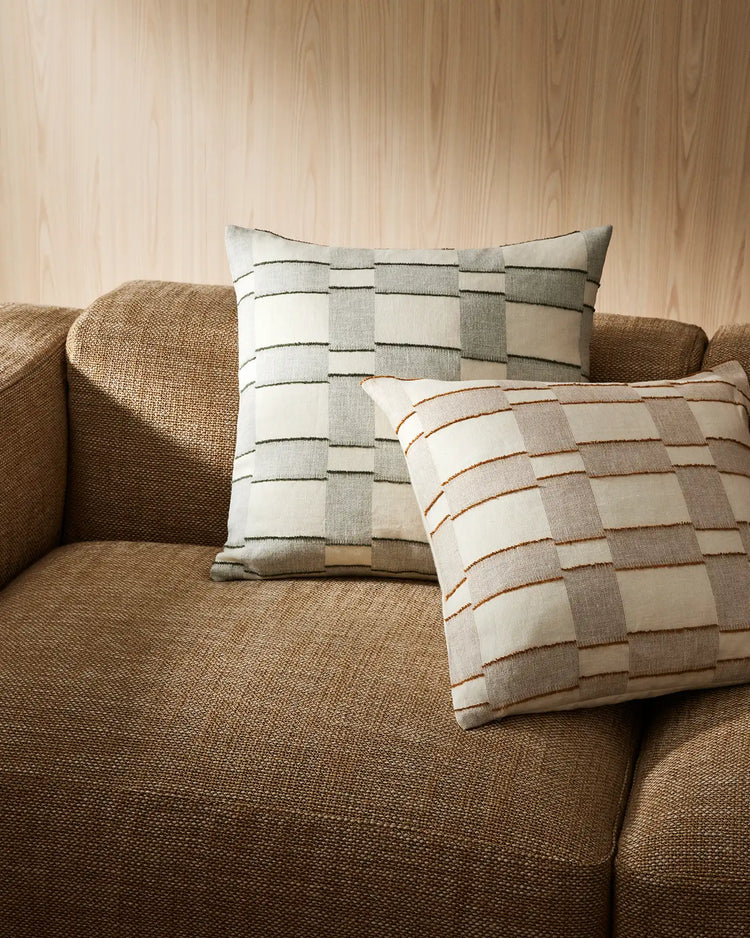 Neutral linen-blend cushions with woven stripe patterns styled on a textured caramel sofa against warm wooden paneling, layering sage, rust and cream tones