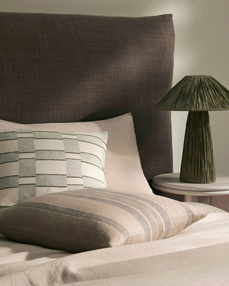 Neutral bedroom with charcoal upholstered headboard, striped linen cushions in cream and sage, and soft geometric bedding layered on white sheets, styled with olive ceramic lamp