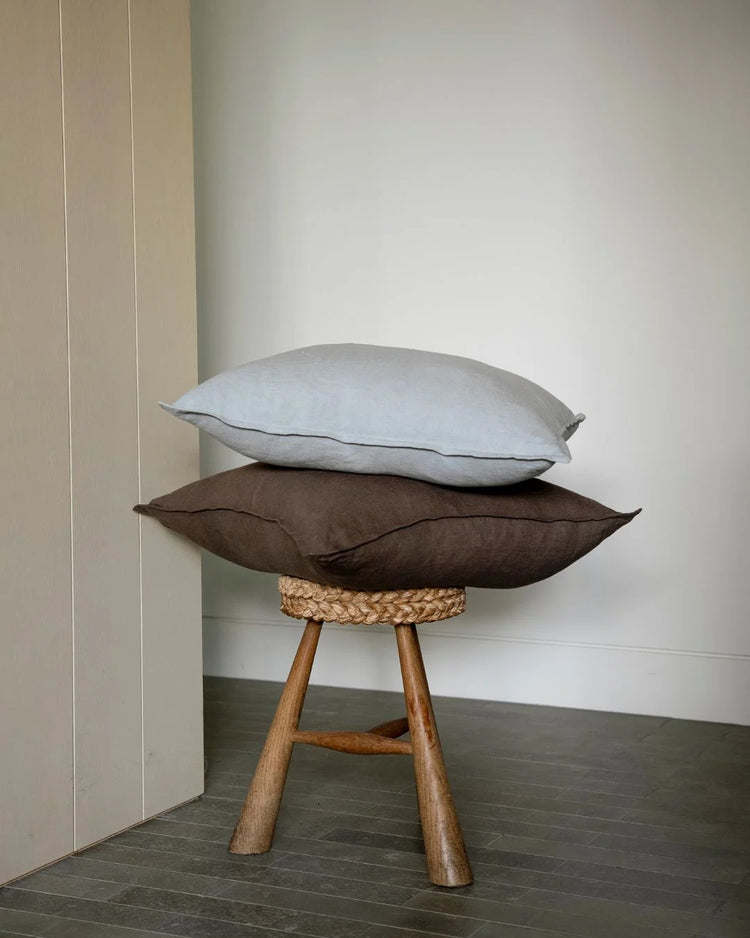 Minimalist interior styling with stacked linen cushions in soft grey and charcoal on wooden tripod stool, neutral palette with natural textures and clean lines