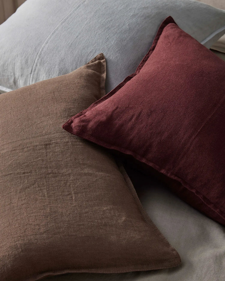 Layered linen cushions in warm mocha and deep garnet tones on a pale grey upholstered chair, creating an earthy, textural palette with natural linen weave detail