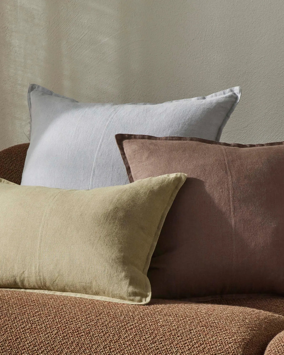 Neutral bedroom styling with Como cushions in sage, mocha and chartreuse linen arranged on a textured caramel throw, against soft beige textured walls.