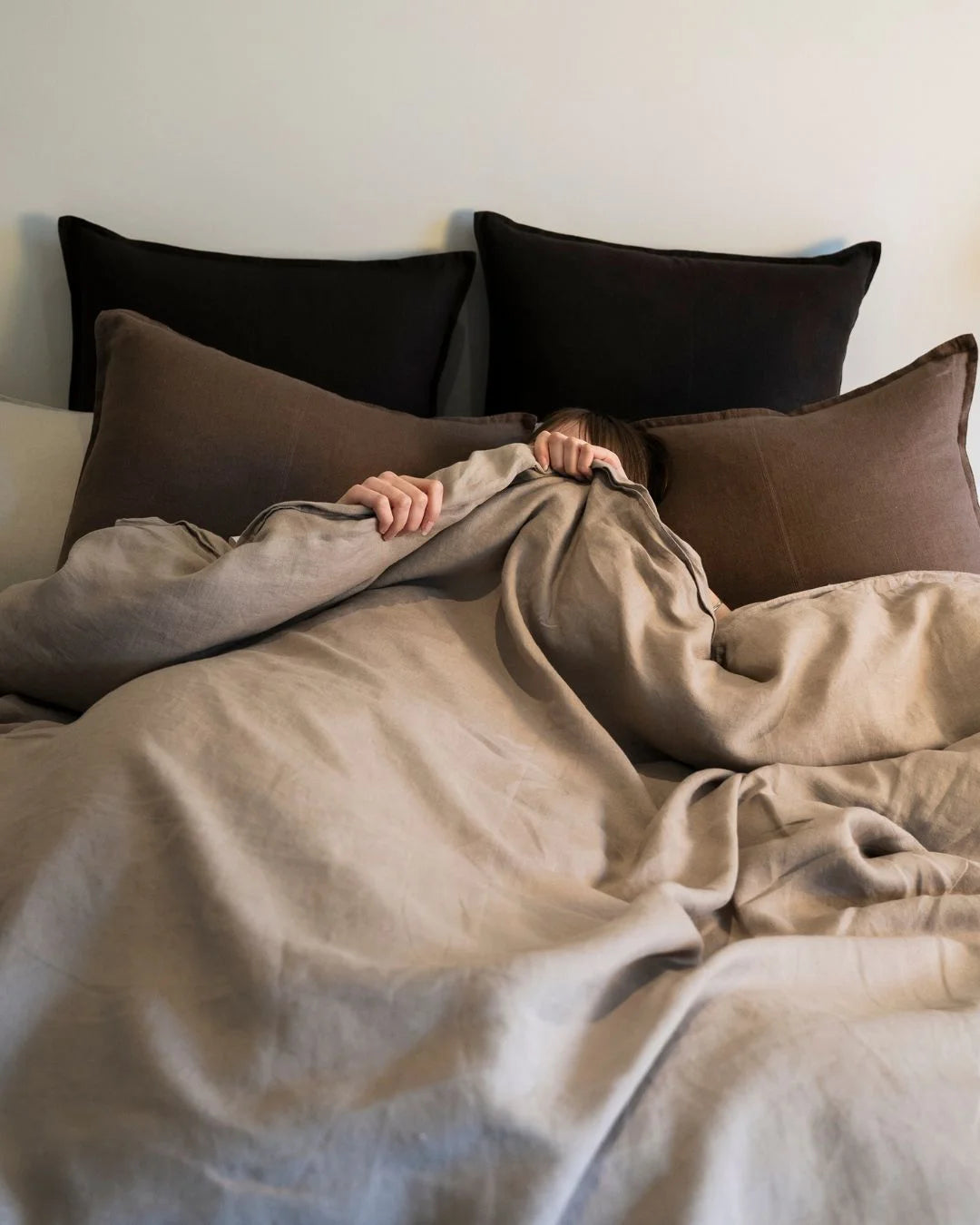 Neutral bedding styled with warm earth tones: pebble linen quilt cover layered with mocha linen cushions and black pillows, creating a minimalist monochromatic bedroom palette.