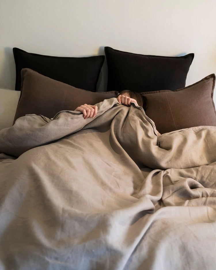 Neutral bedding styled with warm earth tones: pebble linen quilt cover layered with mocha linen cushions and black pillows, creating a minimalist monochromatic bedroom palette.