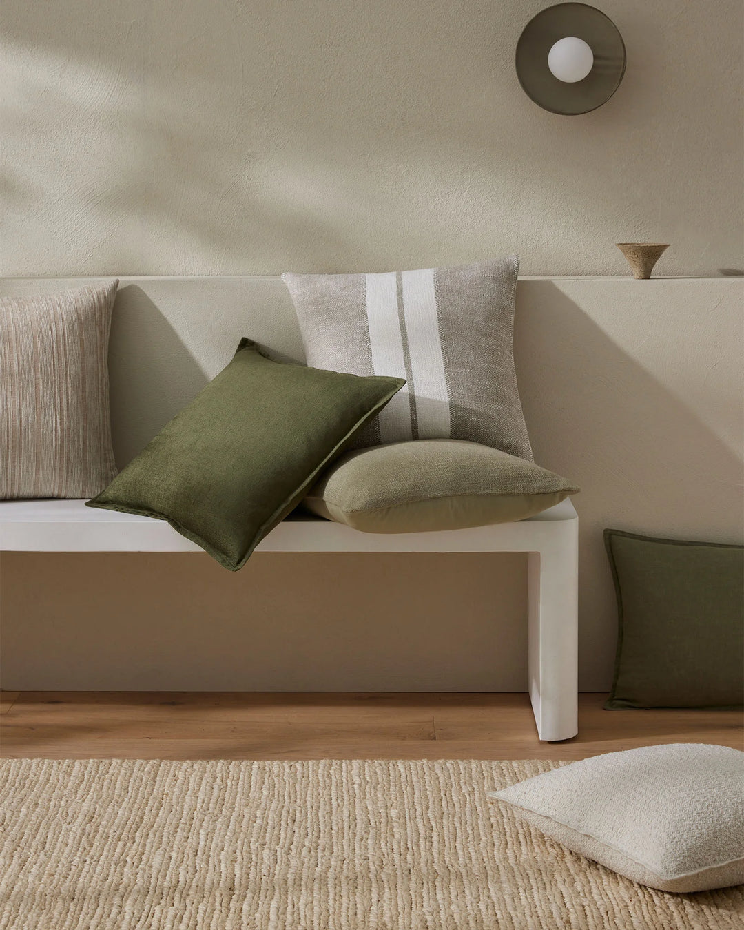 Neutral minimalist bedroom with layered cushions in sage green, olive and cream tones on a beige upholstered bed, striped and textured pillows styled against soft white and natural timber frame