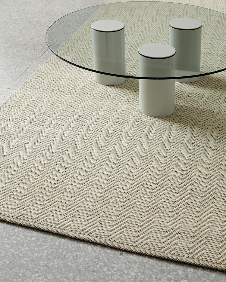 Neutral minimalist living room with cream herringbone wool-cotton rug, glass table, and soft grey cylindrical stools on pale stone floor
