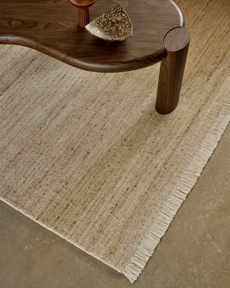 Neutral beige wool rug with fringe edging layered under dark wood table with ceramic accents, warm earthy minimalist interior styling
