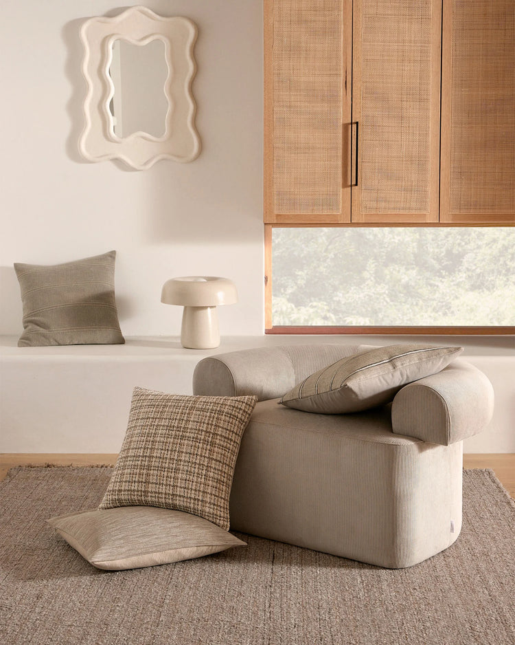 Neutral living room styled with layered cushions in sage, taupe and cream tones on a curved chair, natural jute rug, rattan cabinetry and pale walls creating a serene, textural aesthetic