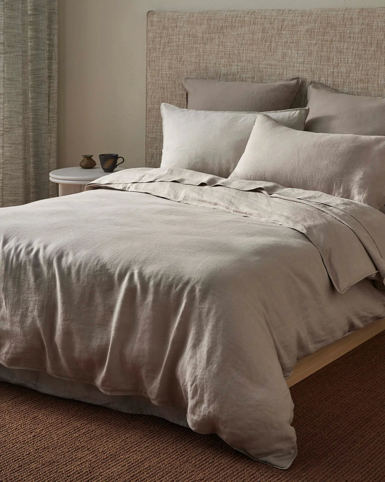 Neutral bedroom with layered linen bedding in warm sand and pebble tones, textured headboard, and earthy minimalist styling with ceramic accessories on wooden nightstand.