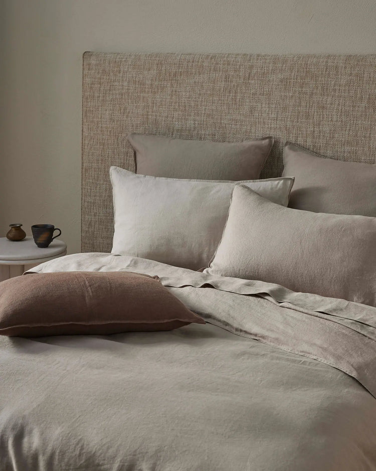 Neutral bedroom scene with layered linen bedding in soft pebble and mocha tones, textured headboard, and warm minimalist styling