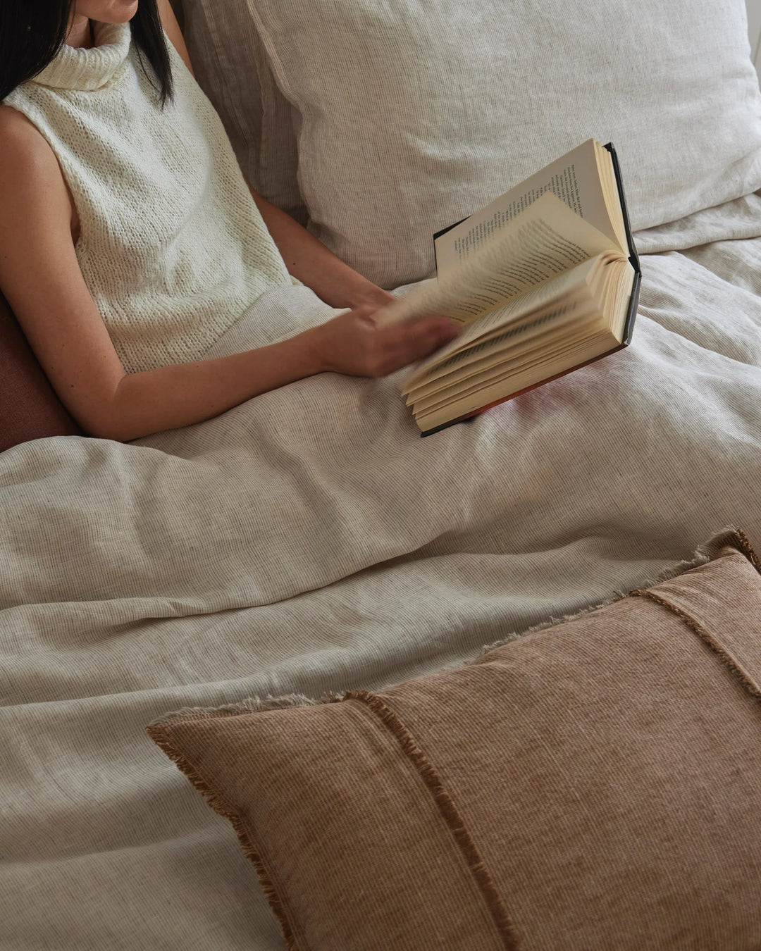 Woman reading on bed layered with neutral linen bedding in cream and taupe tones, featuring textured Sova sheets and Adela rust cushion in natural fibres