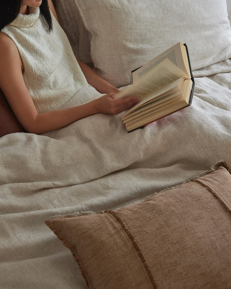 Woman reading on bed layered with neutral linen bedding in cream and taupe tones, featuring textured Sova sheets and Adela rust cushion in natural fibres