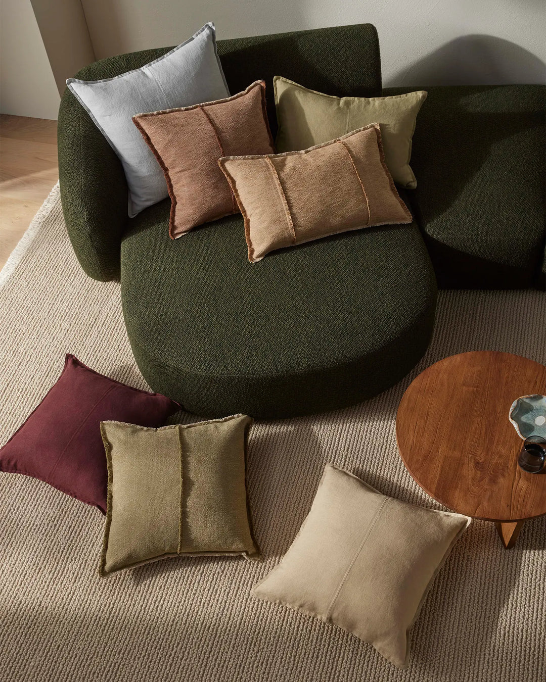 Earthy-toned Adela cushions layered on a forest green curved armchair with warm terracotta, sage and cream accents against natural linen flooring, minimalist styling mood