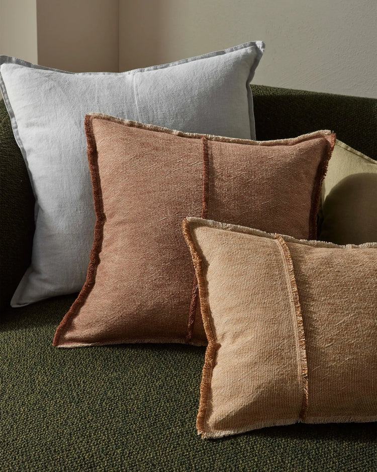 Layered cushions in warm earth tones—rust and terracotta with frayed edges—arranged on a moss-green upholstered sofa with a grey linen pillow, against neutral walls. Warm, textural styling.