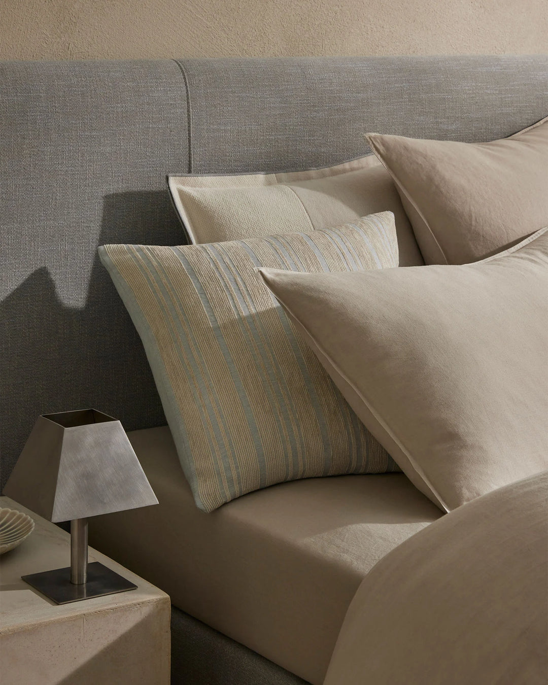 Neutral bedroom styling with layered linen cushions in cream and striped patterns on a grey upholstered bed, soft natural light casting shadows across textured fabrics