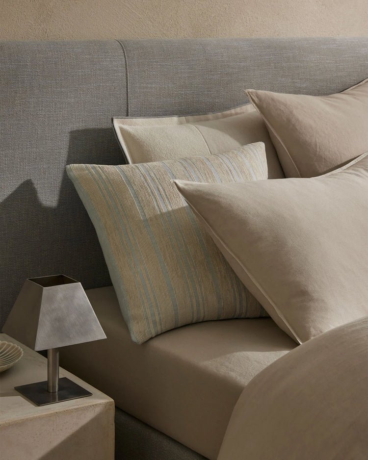 Neutral bedroom styling with layered linen cushions in cream and striped patterns on a grey upholstered bed, soft natural light casting shadows across textured fabrics