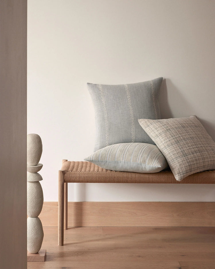 Neutral minimalist bedroom scene with layered linen cushions in soft grey, cream and natural tones on wooden bench, complemented by textured wall art