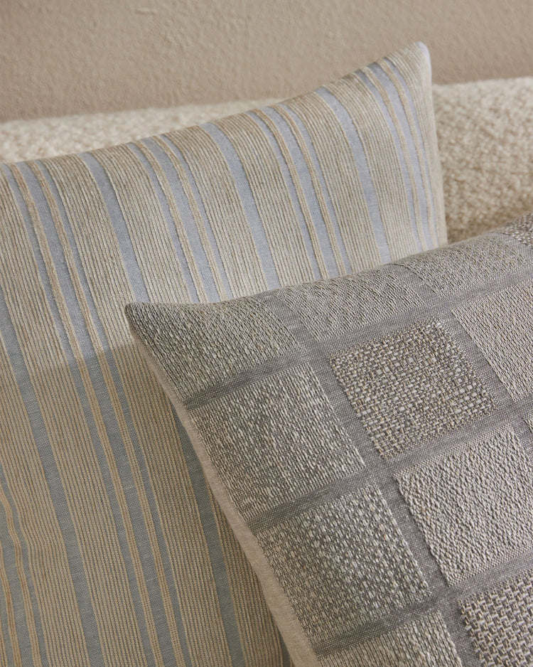 Neutral layered cushions in soft blue, taupe and cream stripes with textured weaves on a cream linen sofa, creating a serene minimalist bedroom aesthetic