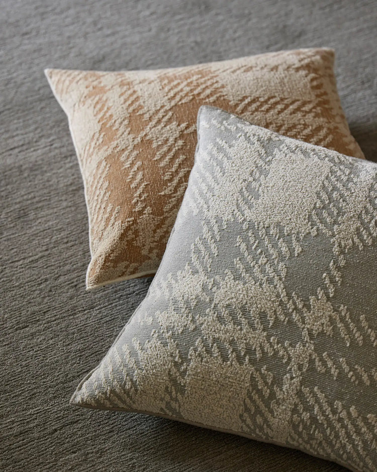 Neutral textured cushions with geometric jacquard weave in warm desert and cool mist tones, styled on grey upholstered surface with layered neutral palette